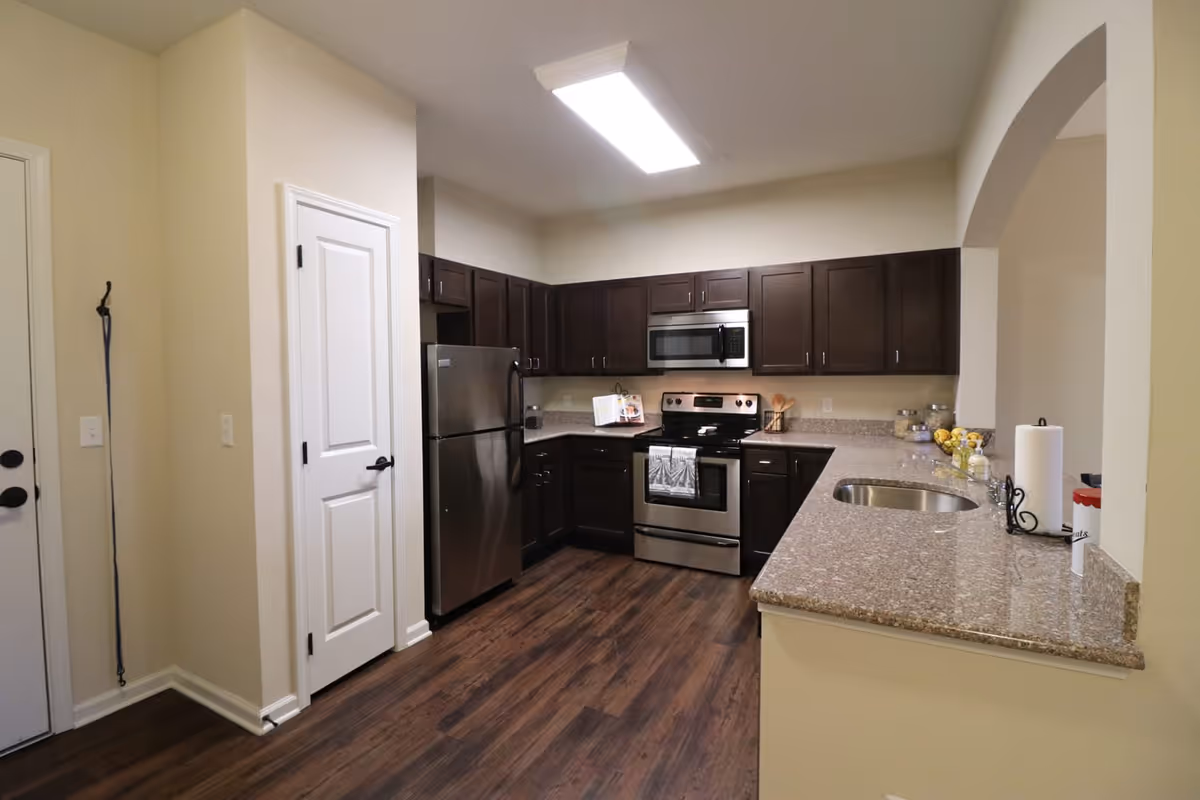 Modern kitchen with dark wood cabinets, stainless steel refrigerator, stove, and microwave. Granite countertops with a sink and paper towel holder. Hardwood flooring and a white door on the left side.