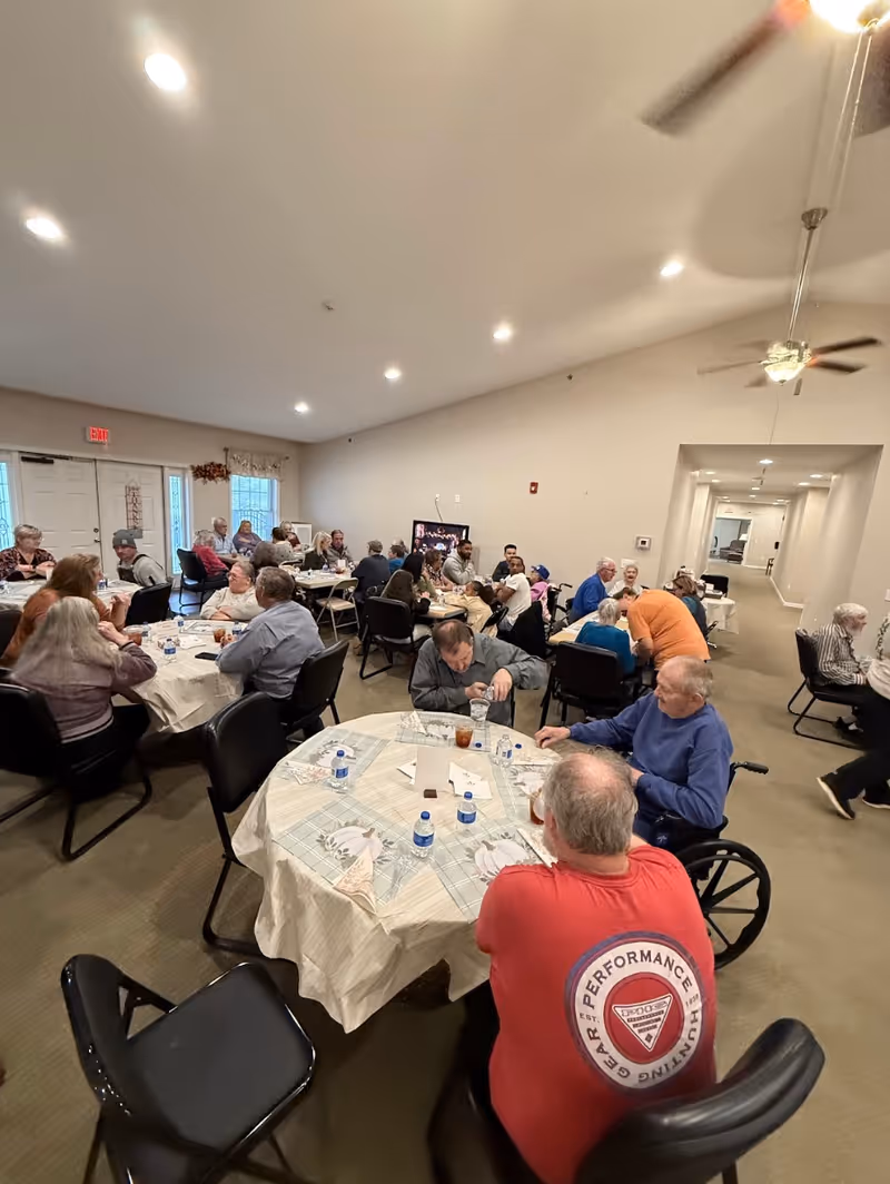 A group of elderly people seated around multiple tables in a large, well-lit room. They appear to be socializing and enjoying a meal or drinks together. The room has beige walls, ceiling fans, and recessed lighting. Some individuals are in wheelchairs, and there are water bottles and glasses on the tables.