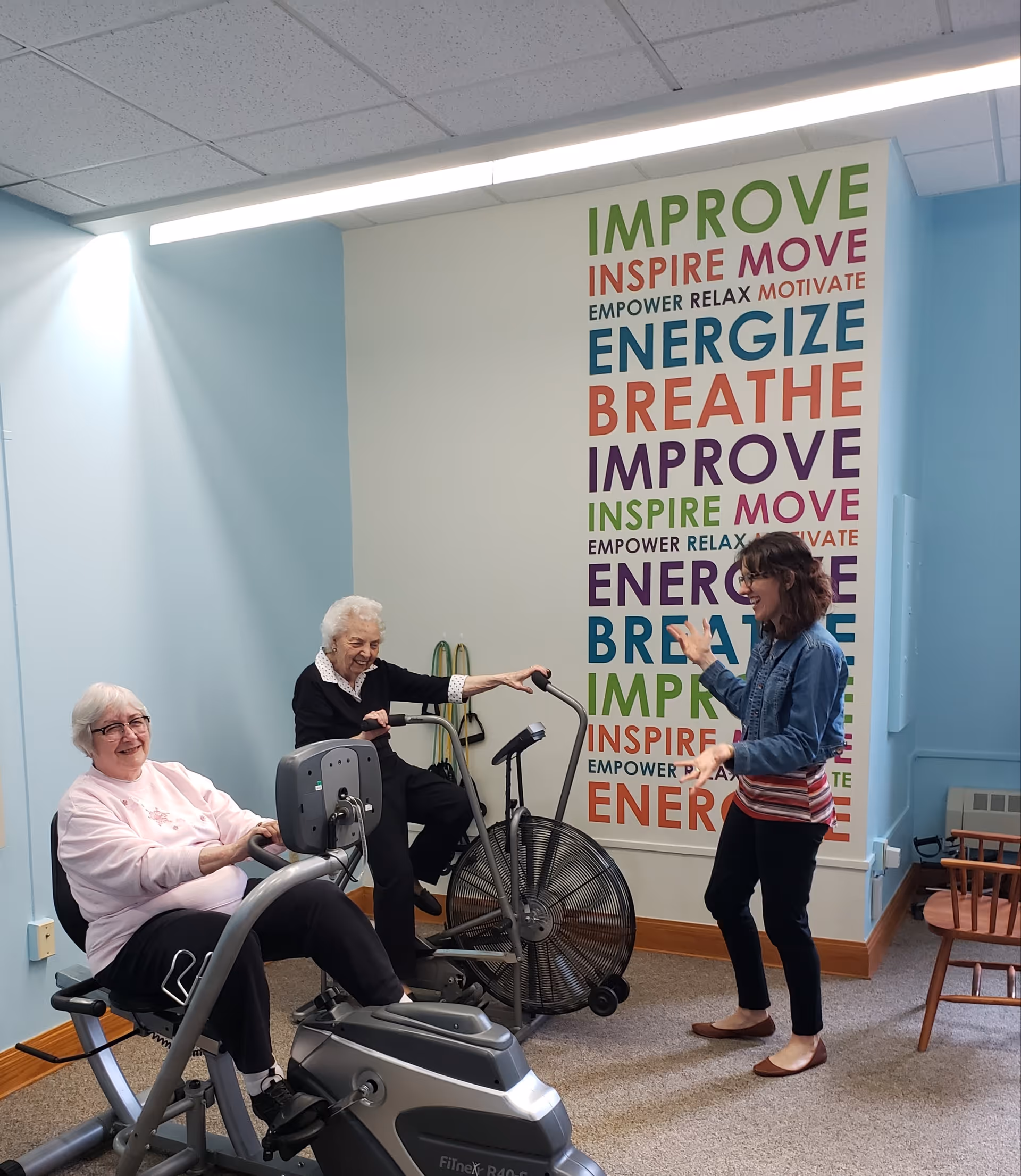Two elderly women exercising on stationary bikes in a light blue room with a motivational word wall behind them. A younger woman stands nearby, engaging with them and smiling. The wall features colorful words like IMPROVE, INSPIRE, MOVE, ENERGIZE, and BREATHE.