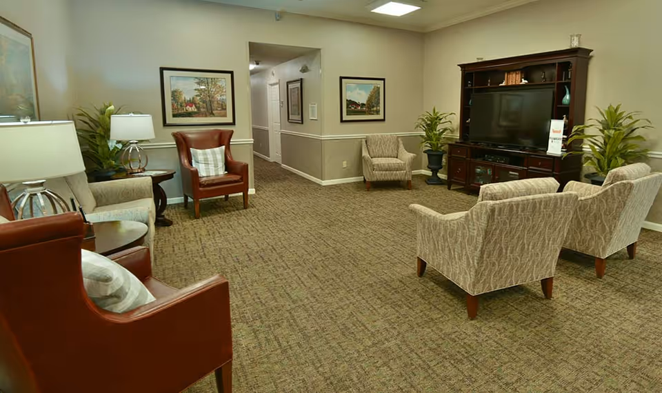 A cozy living room area in an assisted living facility featuring multiple armchairs arranged around a large wooden entertainment center with a flat-screen TV. The room has carpeted flooring, framed artwork on the walls, table lamps, and potted plants adding a touch of greenery.