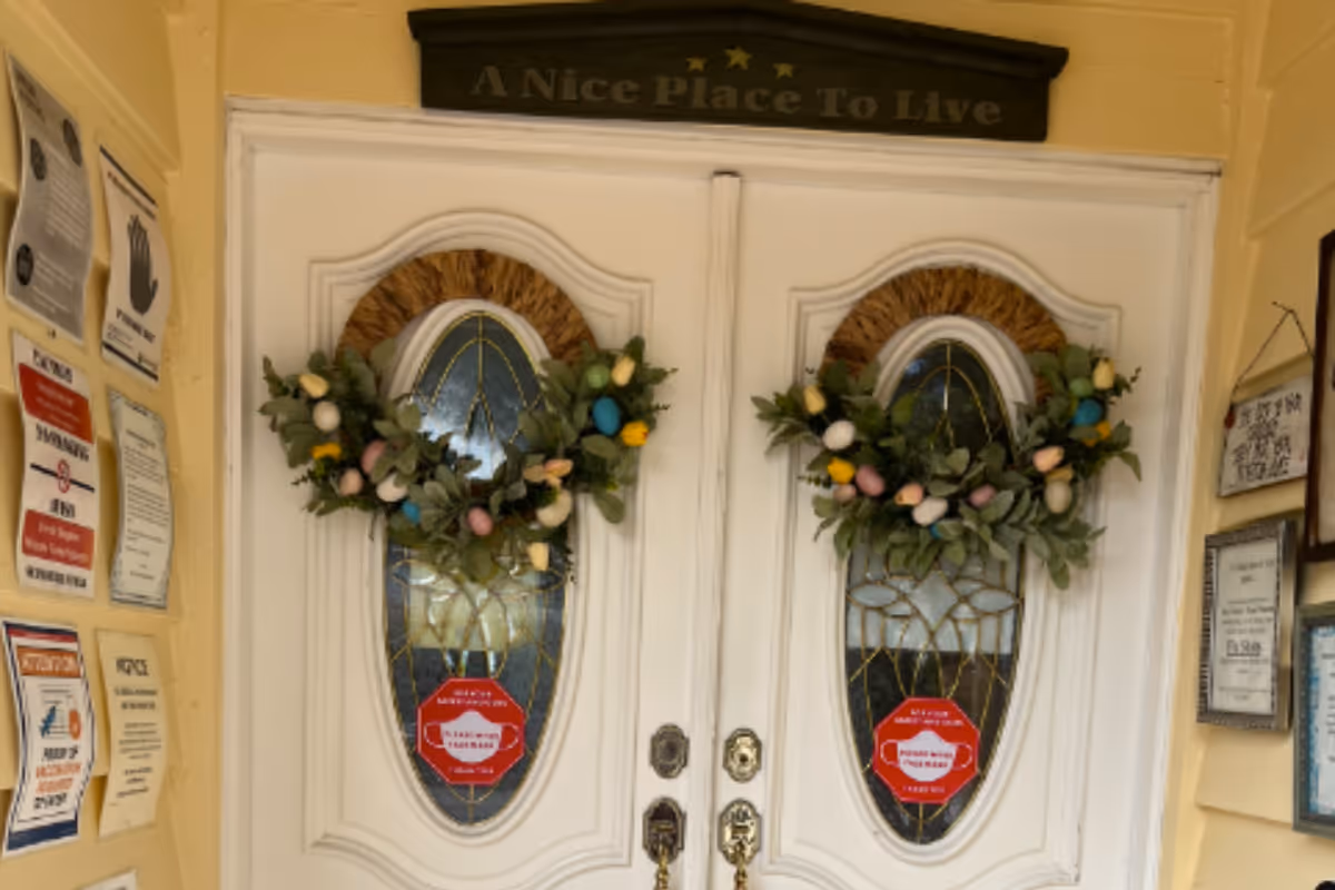 White double front doors with oval stained-glass windows, decorative wreaths, posted notices on either side, and a plaque above reading "A Nice Place To Live".