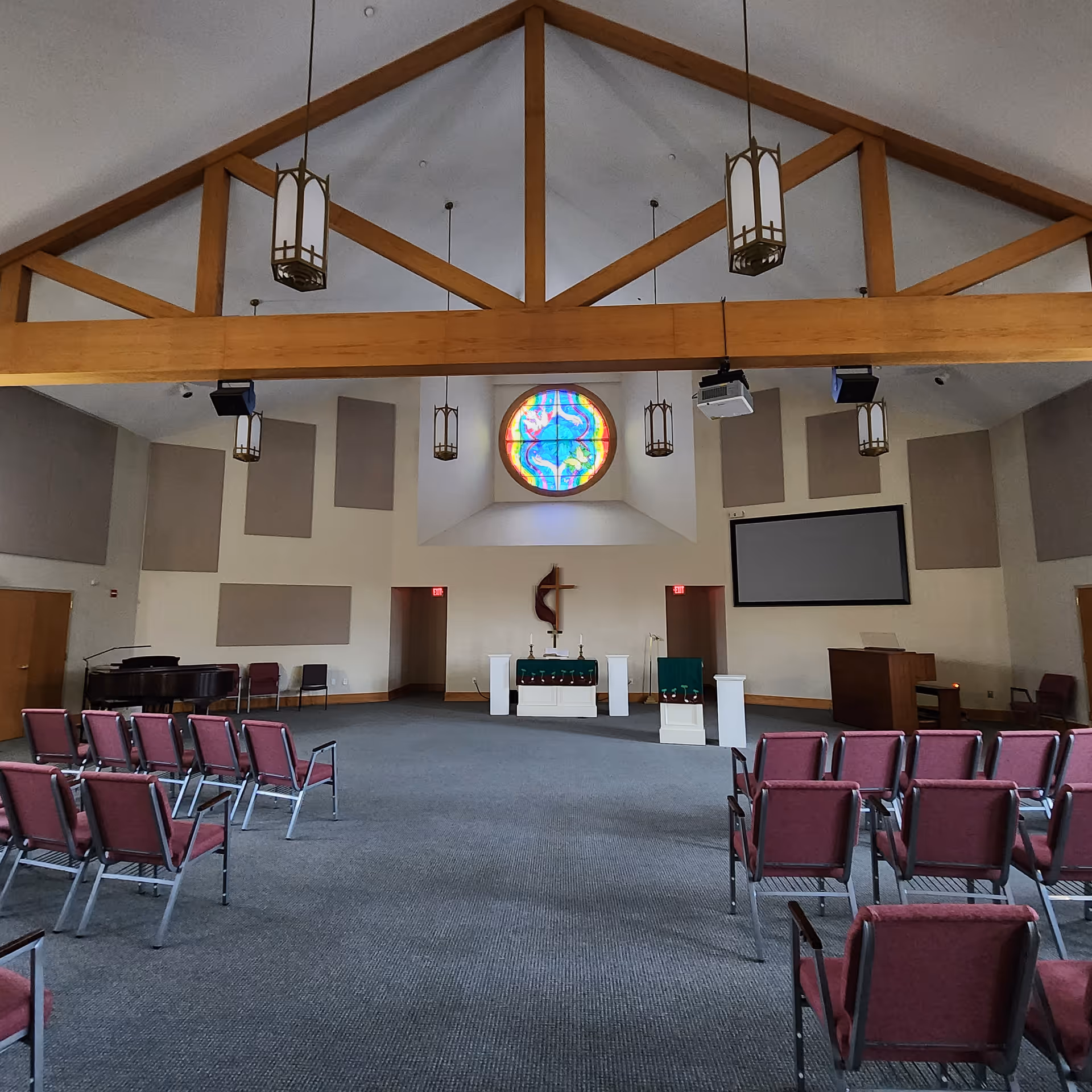 Interior view of a chapel with a high vaulted ceiling supported by wooden beams. There are hanging pendant lights, a stained glass circular window above the altar, a cross mounted on the wall, and rows of red cushioned chairs facing the altar. A piano and a lectern are visible on the sides.