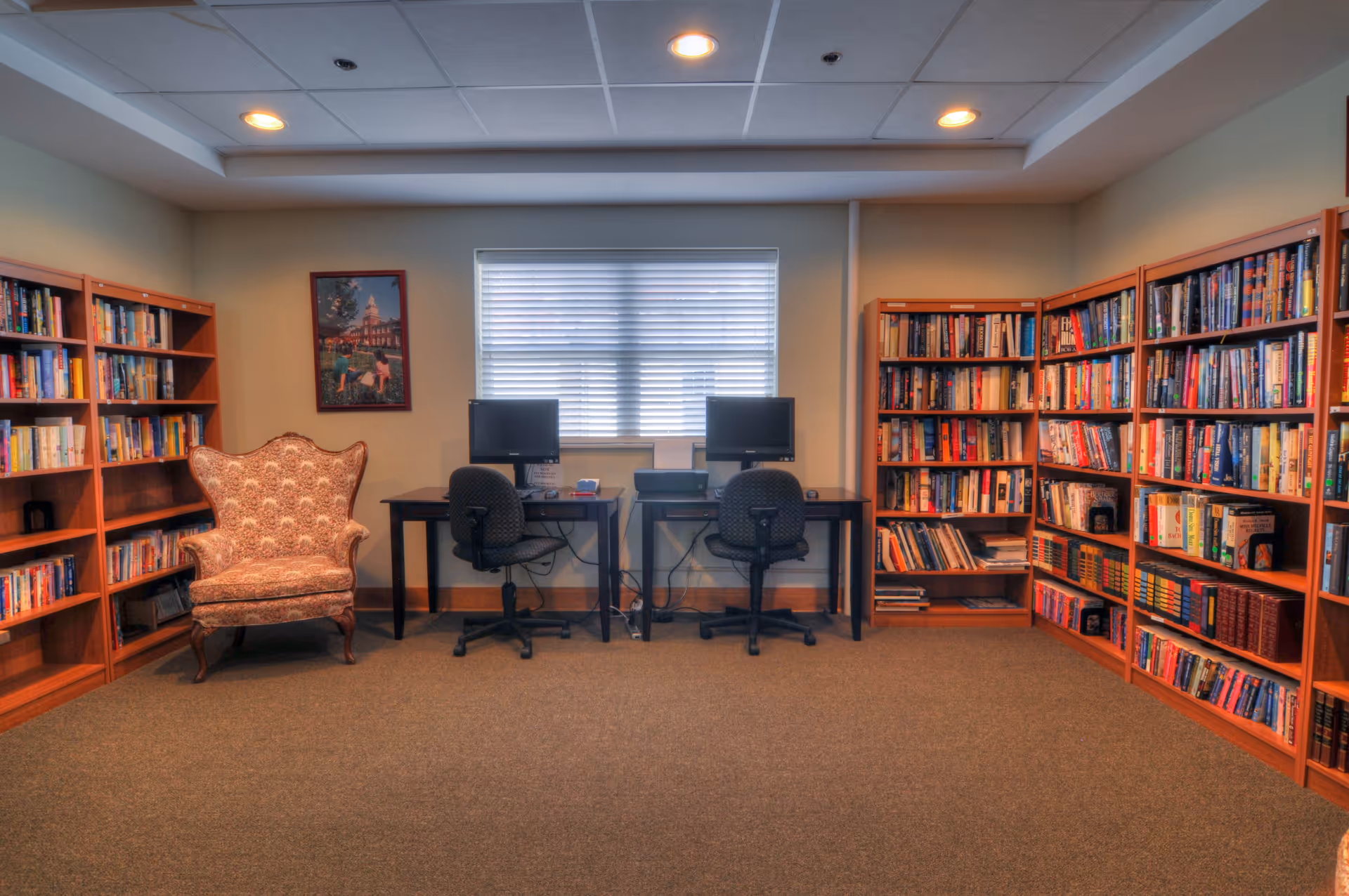 A quiet room with bookshelves filled with books along two walls, a patterned armchair, and two computer workstations with black office chairs in front of a window with blinds.