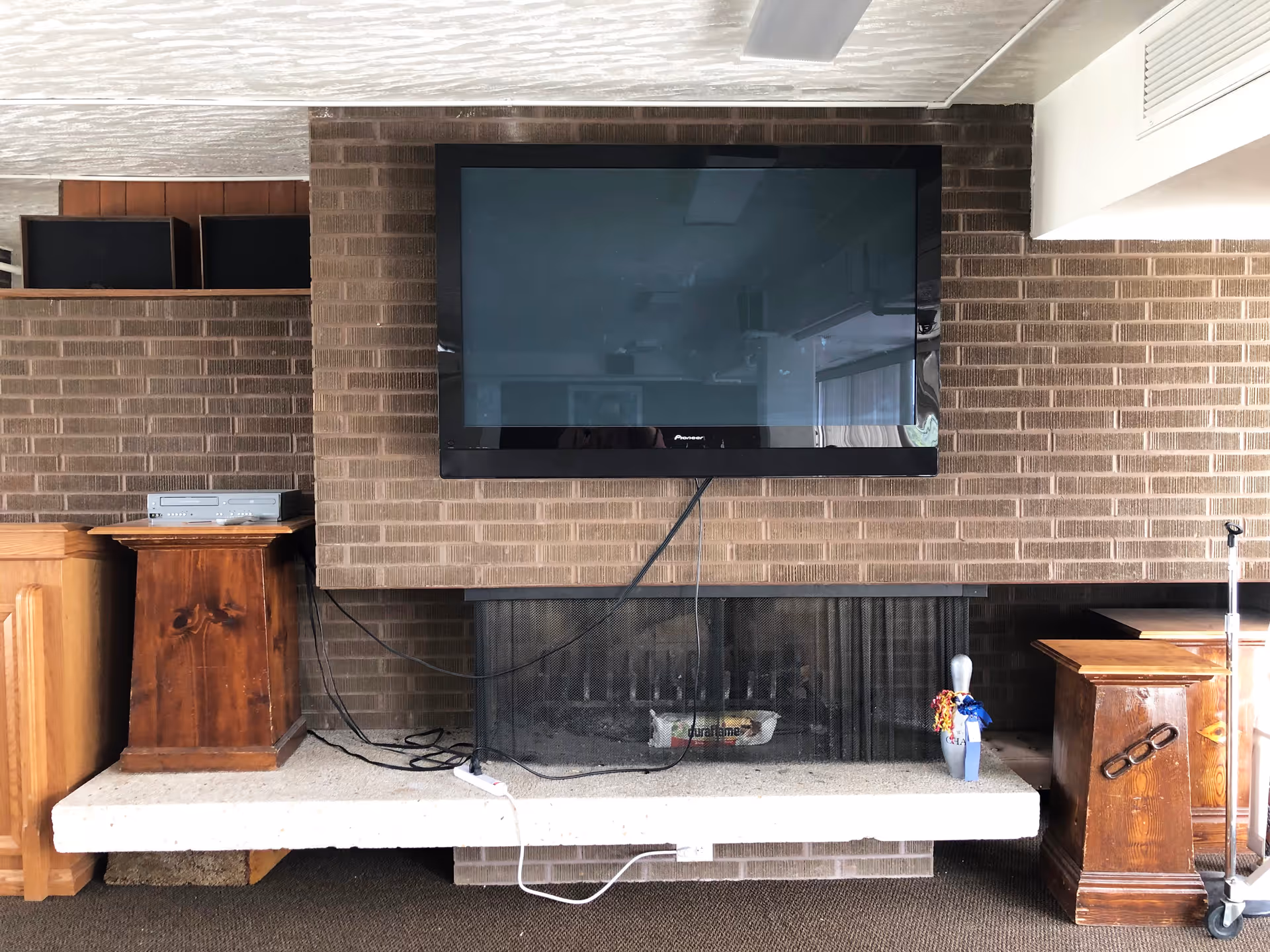A living room area with a large flat-screen Pioneer TV mounted on a brown brick wall above a fireplace. The fireplace has a black mesh screen and a white stone hearth. On the hearth, there is a small bowling pin decorated with colorful ribbons. To the left and right of the fireplace are wooden pedestals, and there is a silver electronic device with a remote on the left pedestal. The floor is carpeted in a dark color.