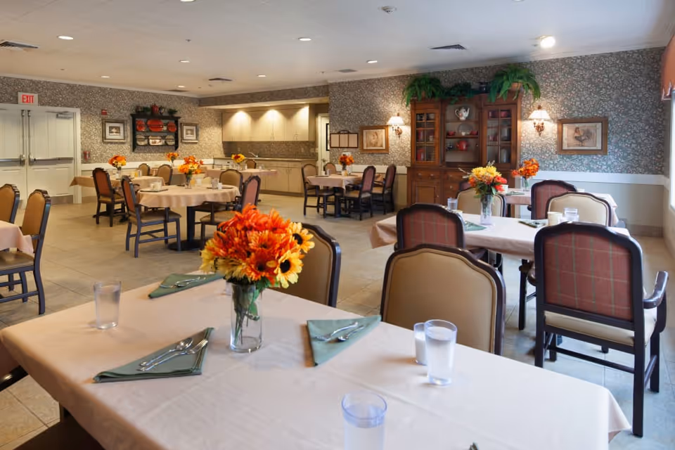 Dining room with tables set with napkins, water glasses and orange flower centerpieces, chairs, and a wooden cabinet against patterned wallpaper.