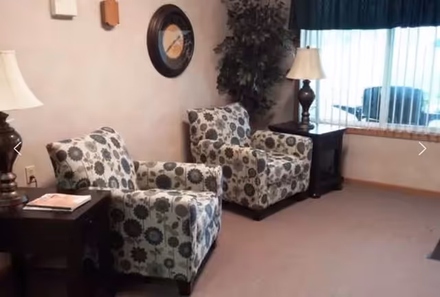 Two patterned armchairs with side tables and lamps in a seating area by a window with vertical blinds.