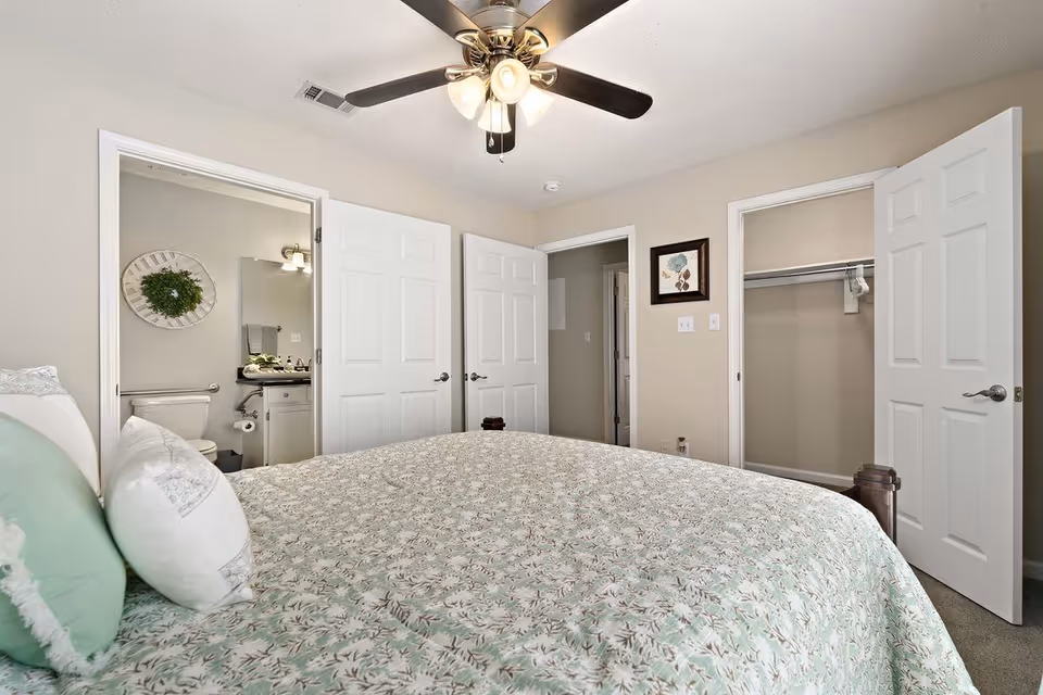Bedroom with a bed covered in a green and white floral bedspread, two pillows, a ceiling fan with lights, an open door leading to a bathroom with a toilet and sink, and another open door revealing an empty closet.