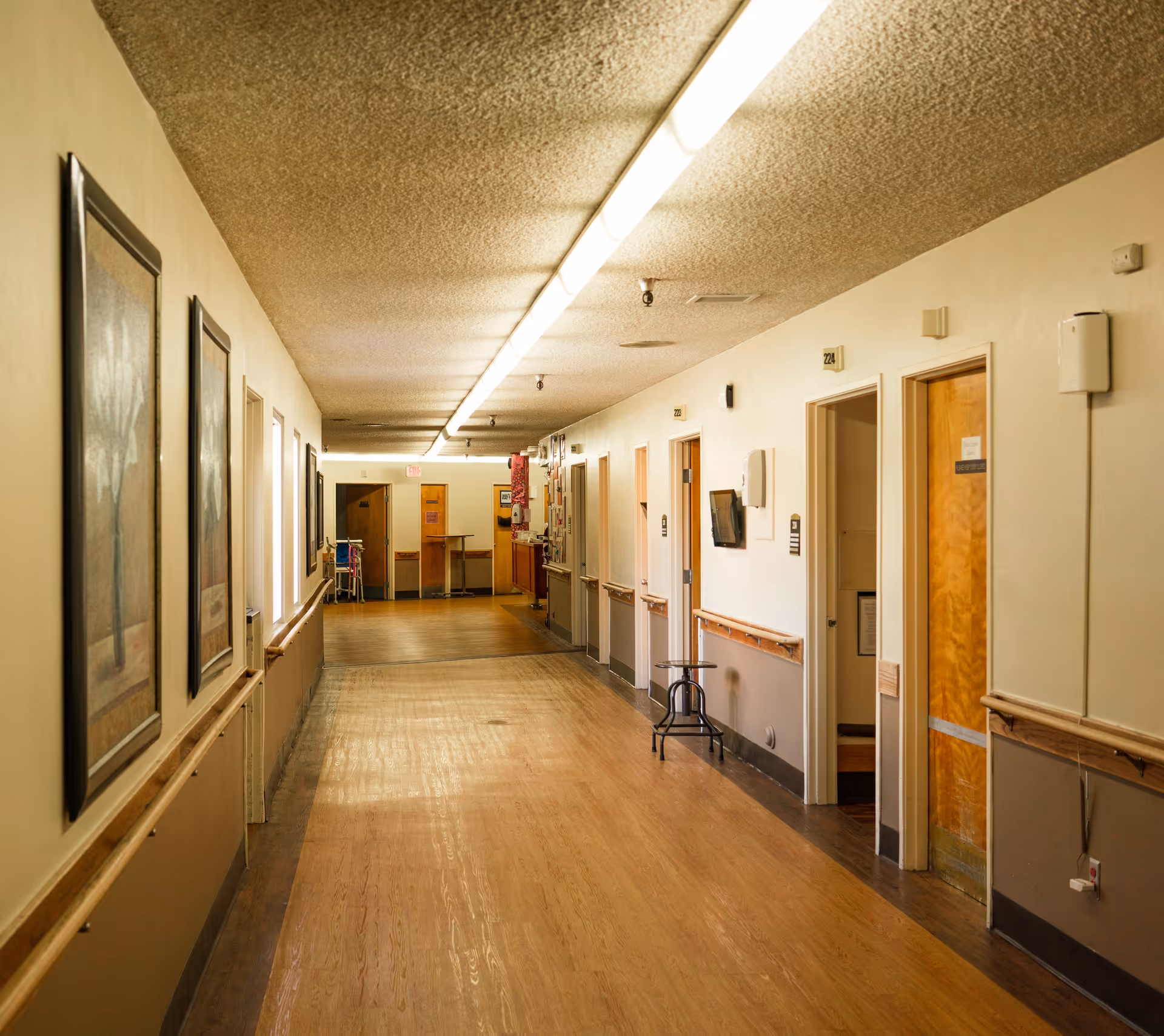 A long, well-lit hallway in a healthcare facility with wooden floors and beige walls. Several doors line the right side of the hallway, some open, with handrails along the walls. There are framed pictures on the left wall and a stool near one of the doors. The ceiling has a textured finish with fluorescent lighting running down the center.