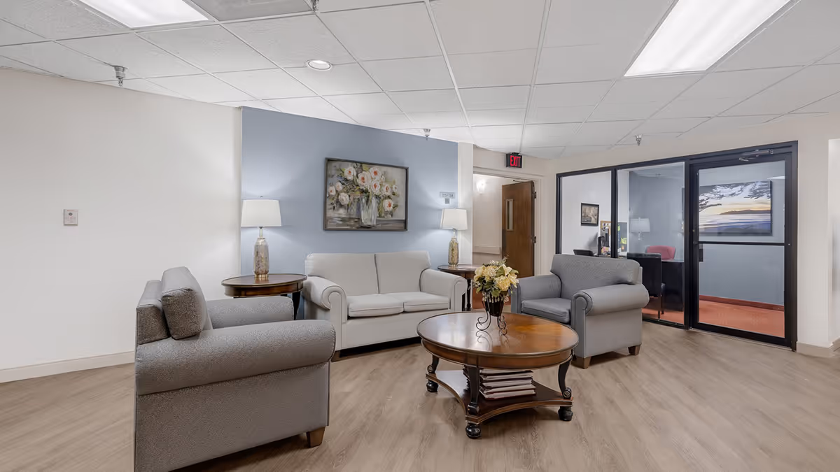 A cozy senior living facility common area with two gray armchairs and a light gray sofa arranged around a round wooden coffee table with a flower vase on top. Two table lamps are placed on side tables next to the sofa, and a painting of white flowers hangs on a blue accent wall. There is a door with an exit sign above it and glass walls showing an adjacent room with a desk and chairs.