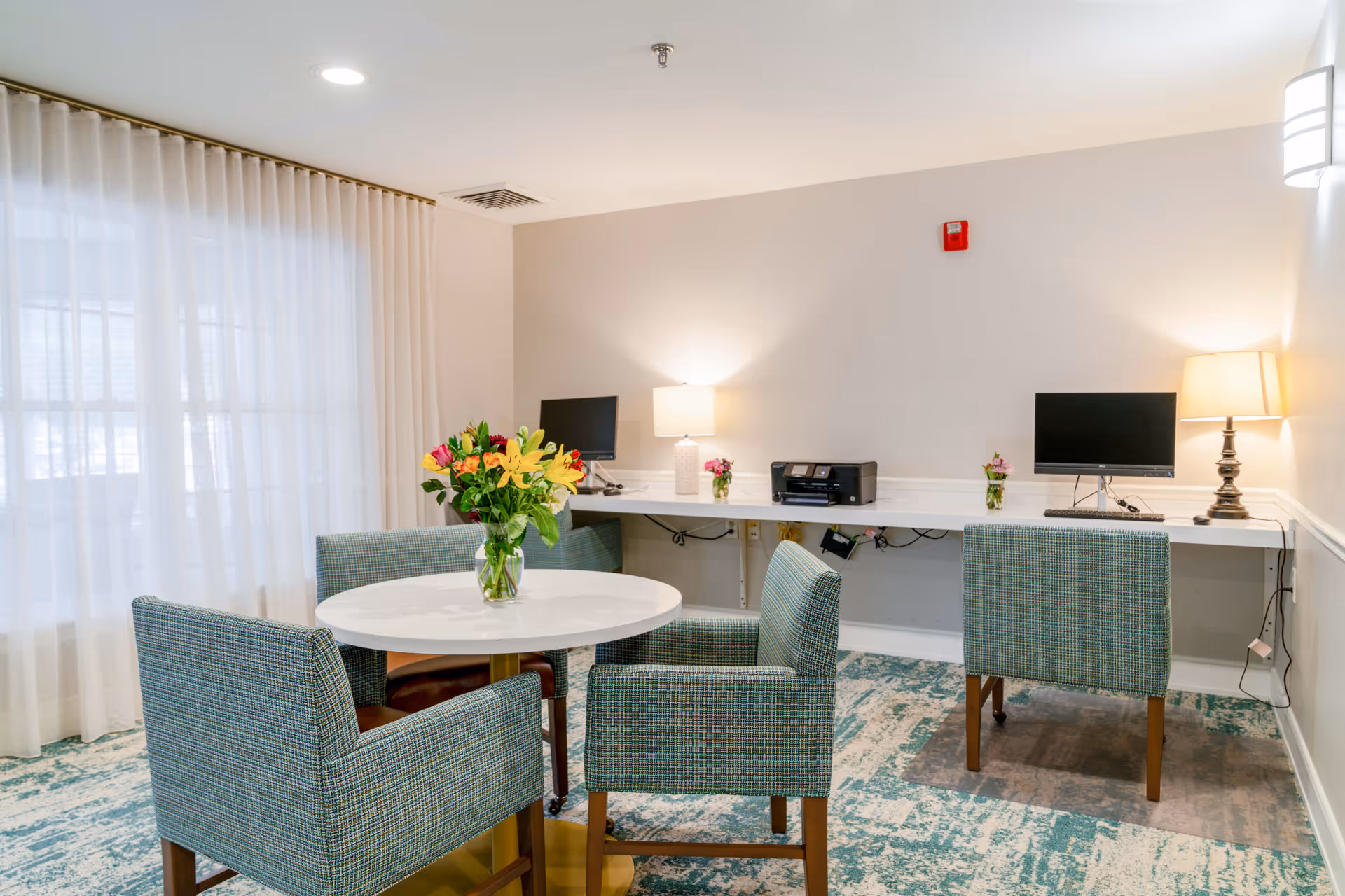 A bright room with a round white table surrounded by four upholstered chairs with a blue and green pattern. A vase with colorful flowers is placed on the table. Against the wall, there is a long white desk with two computer monitors, a printer, and two table lamps providing warm light. The floor has a patterned carpet in shades of blue and beige, and sheer white curtains cover the window on the left side.