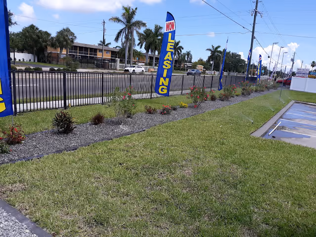 Outdoor view of a grassy area with a landscaped flower bed along a black metal fence. Several blue and yellow flags with the text 'NOW LEASING' are placed along the fence. Palm trees and a road with cars are visible in the background under a partly cloudy sky.