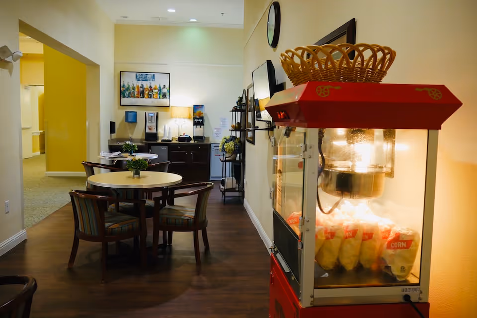 Interior view of a senior living facility common area with a popcorn machine in the foreground on the right, a round table with four chairs in the middle, and a beverage station with a water dispenser and coffee machine in the background. The walls are light-colored, and there is a framed picture of soda bottles on the wall.