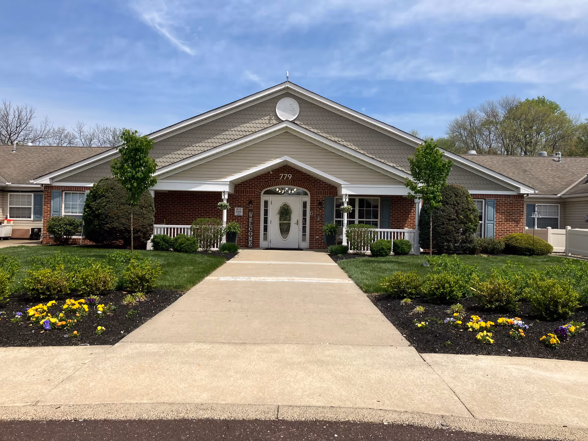 Front exterior view of a single-story brick and siding building with a peaked roof, a central entrance with a white door and sidelights, flanked by two small trees and landscaped flower beds with yellow and purple flowers under a partly cloudy sky.