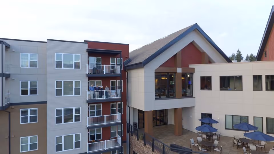 Exterior view of a multi-story senior living facility with balconies and a patio area featuring tables and umbrellas. Several people are standing on one of the balconies. The building has a modern design with a mix of beige, brown, and red siding and large windows.