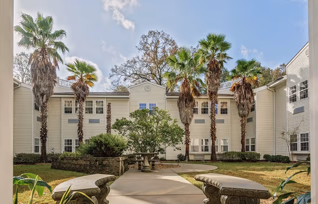 Outdoor courtyard area at Sodalis Tallahassee Assisted Living featuring a paved walkway, stone benches, palm trees, and a two-story building with multiple windows under a partly cloudy sky.