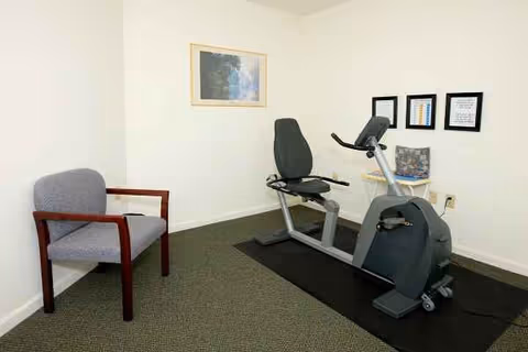 Small interior room with exercise bike, chair, framed certificates, and wall art.
