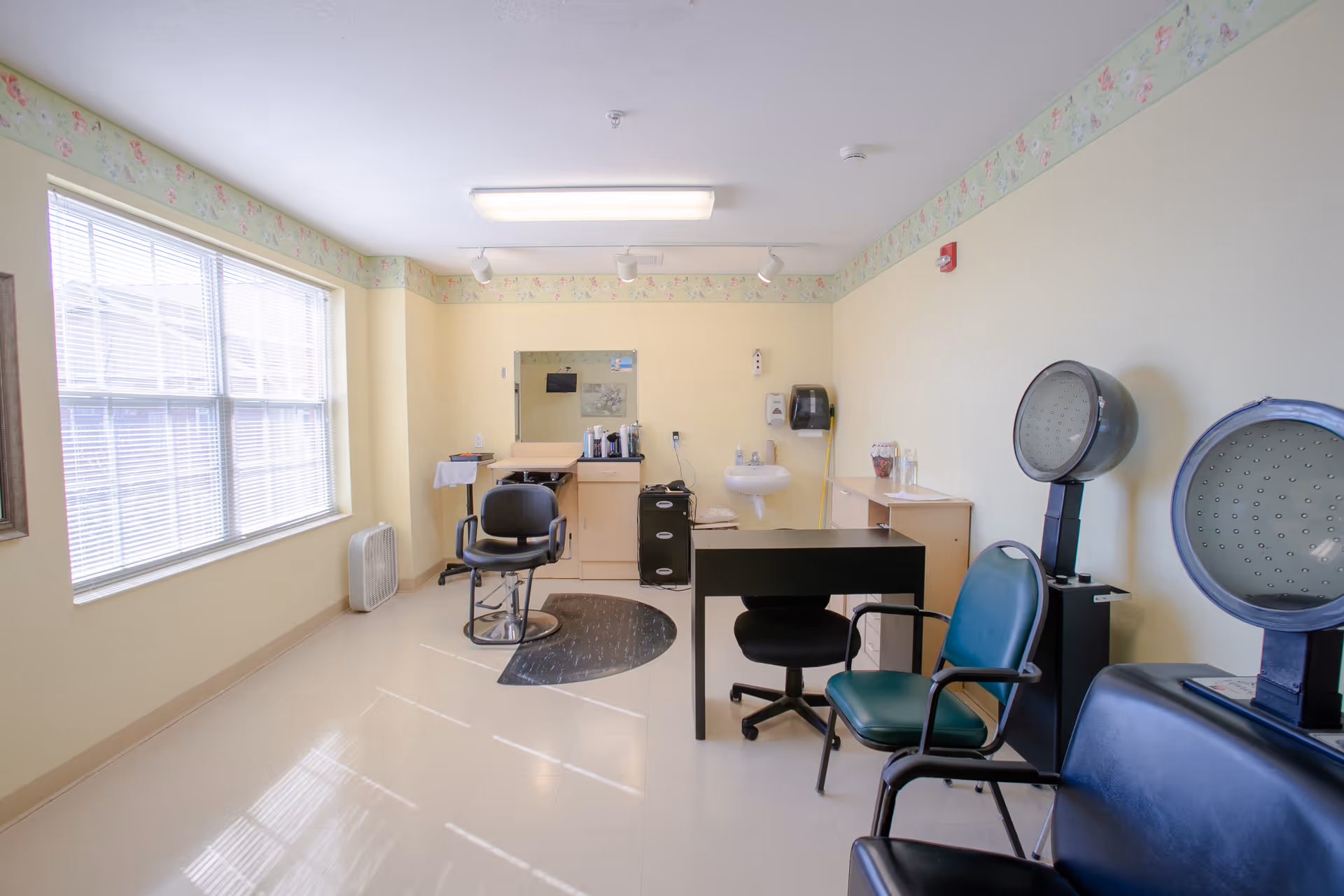 A bright room with large windows letting in natural light, featuring salon chairs, hair drying stations, a small desk with a chair, a sink, and cabinets. The walls are light yellow with a floral border near the ceiling.