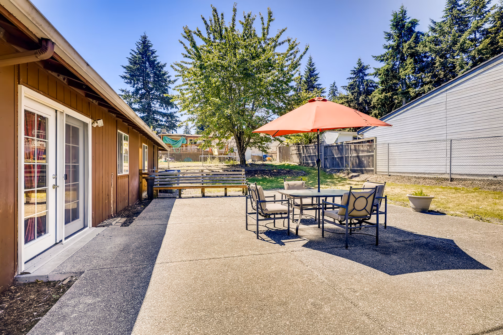 Outdoor patio area at Evergreen Senior Care with a round table and four cushioned chairs under a red umbrella, a wooden bench along the building wall, a tree providing shade, and a fenced yard with grass and tall evergreen trees in the background.