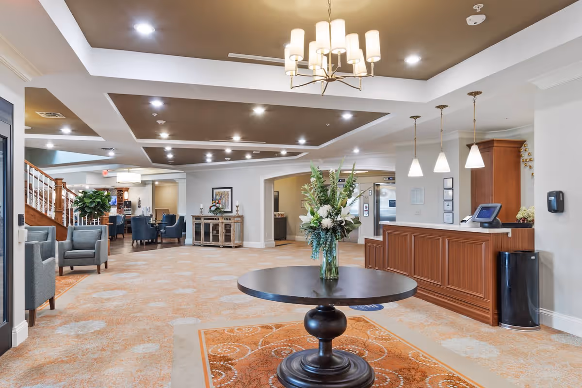 Spacious and well-lit senior living facility lobby with a round table holding a floral arrangement in the center, a wooden reception desk with pendant lights on the right, comfortable seating areas with armchairs and tables on the left, and a staircase leading upstairs in the background.