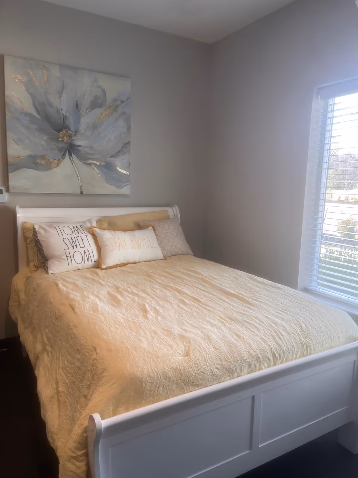Bright bedroom with a white bed dressed in a pale yellow quilt and decorative pillows (one reading "HOME SWEET HOME") beneath a large floral painting beside a window with blinds.