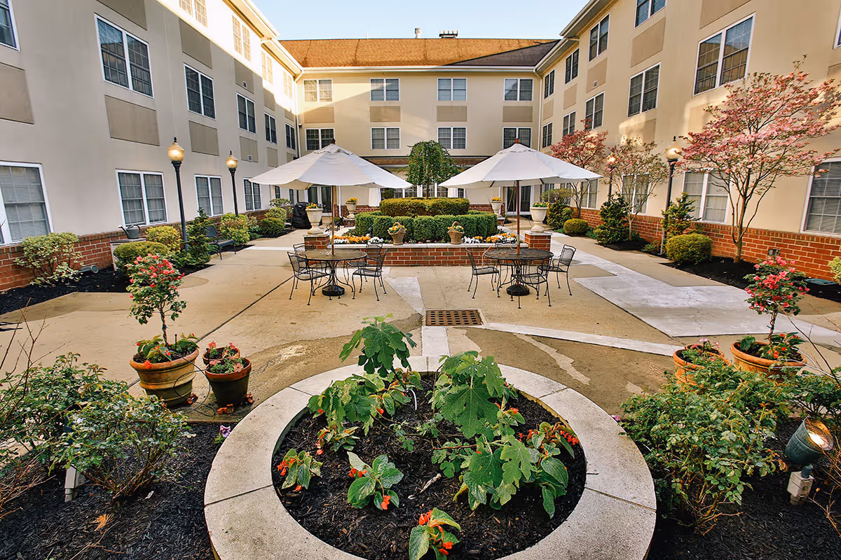 Outdoor courtyard area of a senior living facility with a circular flower bed in the foreground, surrounded by potted plants and shrubs. There are two tables with umbrellas and chairs on a paved patio, with a three-story beige building with many windows surrounding the courtyard. Small trees and bushes line the edges of the courtyard.