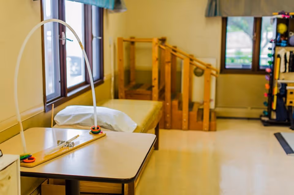 A therapy room with a table featuring a ring toss game, a padded bench with a pillow, wooden stairs with handrails for physical therapy, and exercise equipment near the windows.