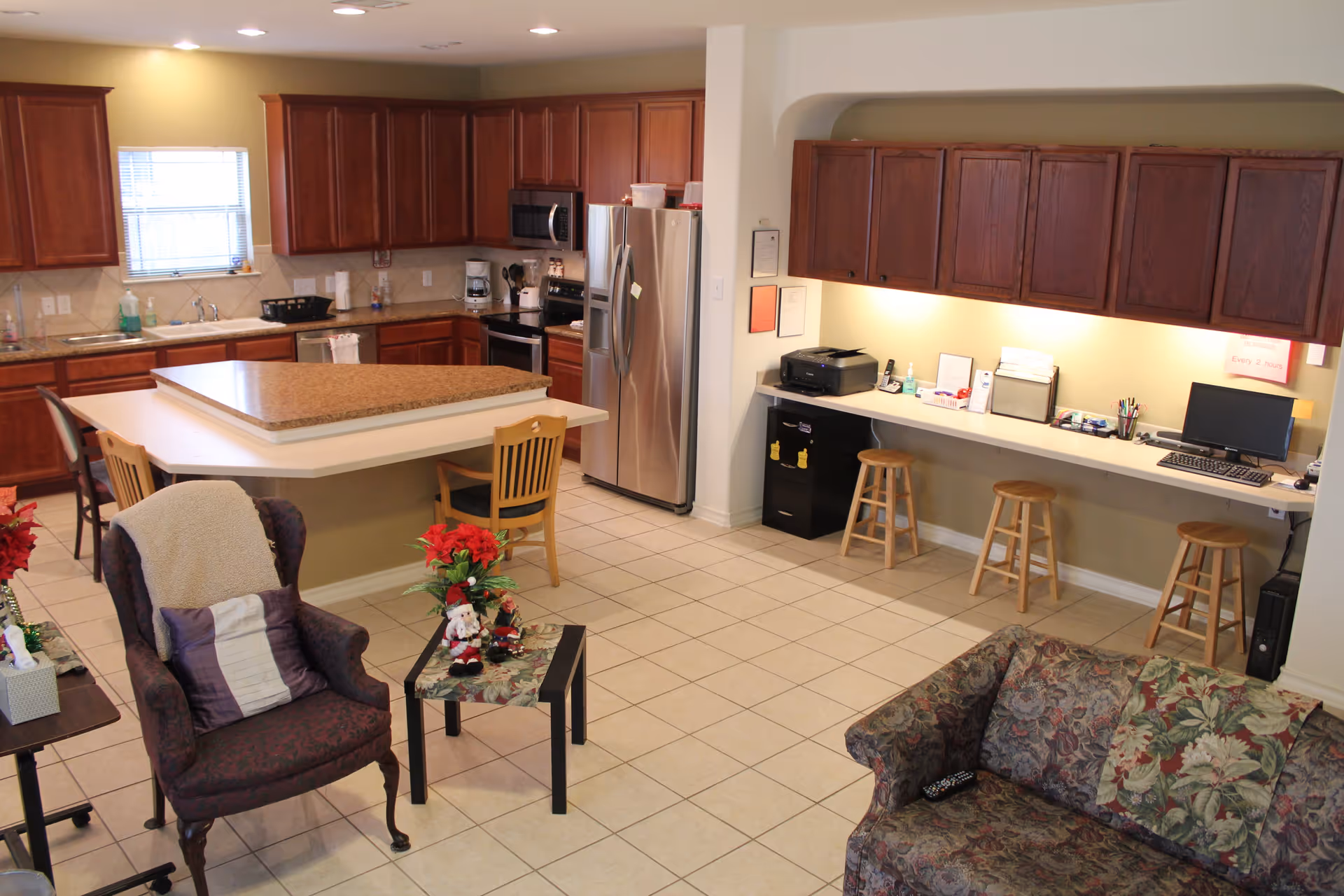 Interior view of a senior living facility common area featuring a kitchen with wooden cabinets, a central island, stainless steel refrigerator, and appliances. Adjacent to the kitchen is a workspace with a long counter, three wooden stools, a computer, printer, and office supplies. In the foreground, there is a floral-patterned sofa, an upholstered armchair with a blanket and pillow, and a small table with holiday decorations.
