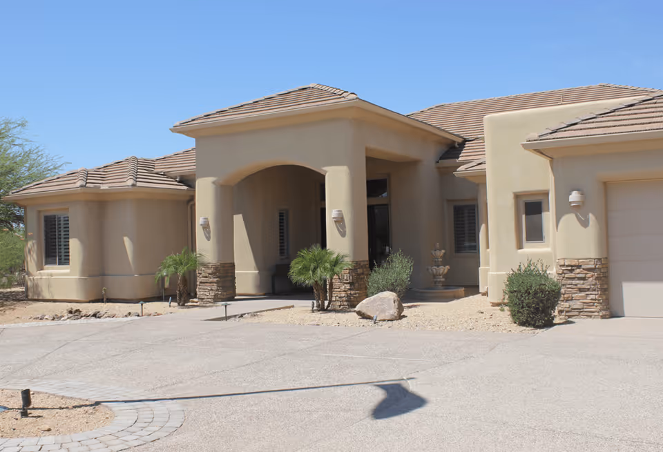 Exterior front view of a single-story beige stucco building with a tiled roof, featuring an arched entrance supported by columns with stone bases, small palm plants, and desert landscaping under a clear blue sky.