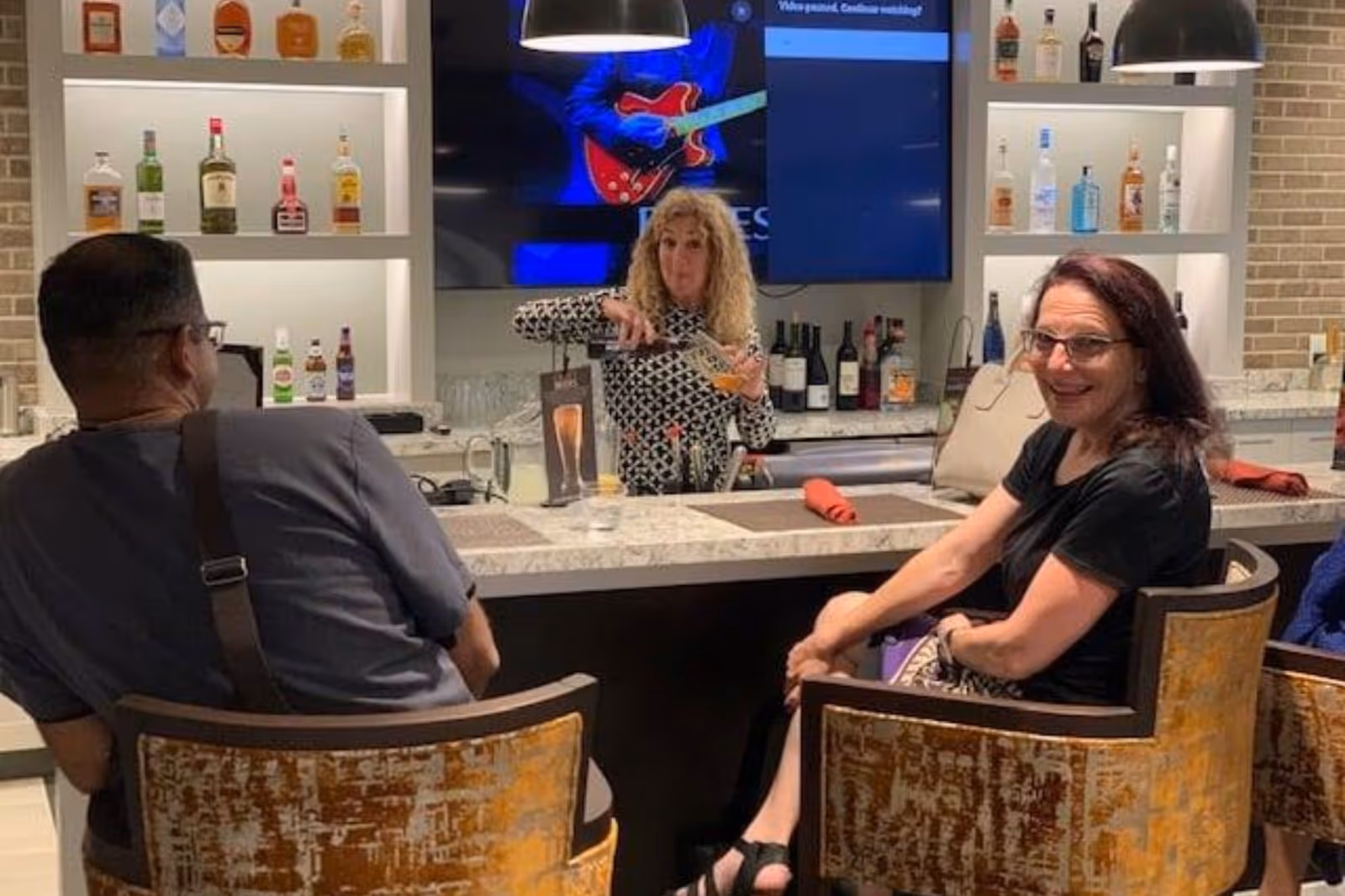 Three people sit at a marble bar while a server pours a drink in a well-lit lounge with bottles and a TV behind the counter.