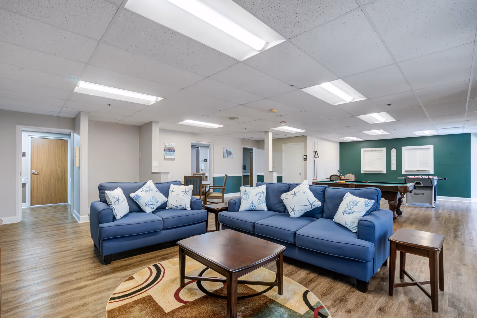 A spacious common area in a senior living facility featuring two blue sofas with decorative pillows arranged around a wooden coffee table on a patterned rug. The room has wood flooring, fluorescent ceiling lights, and a green accent wall in the background with two windows. There is a pool table and an air hockey table near the back wall, and a small dining table with chairs is visible near the center. Several doorways lead to other rooms.