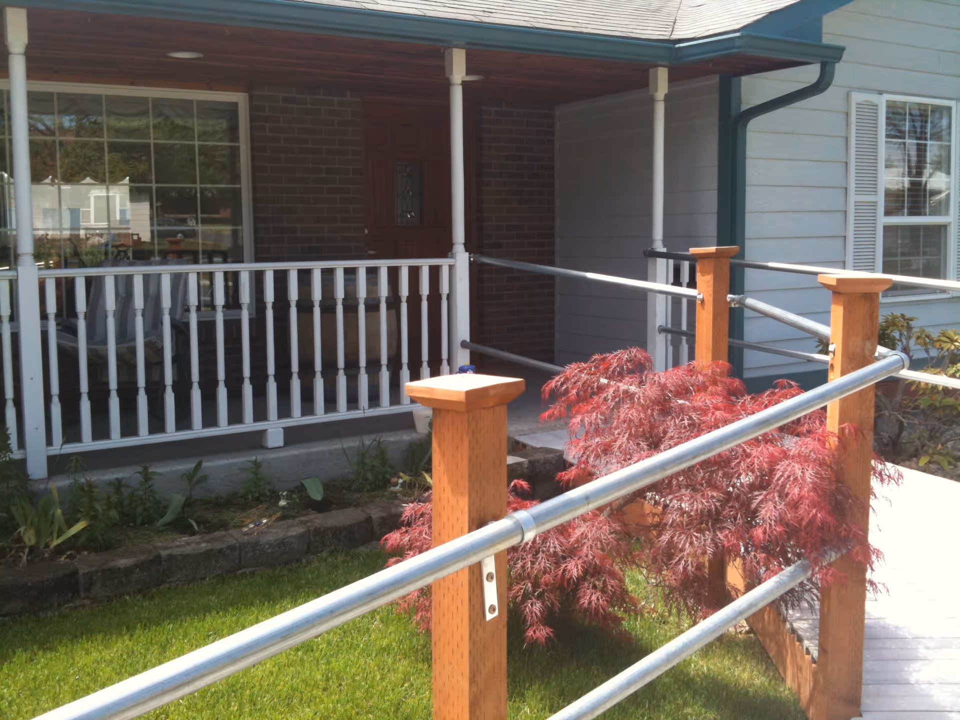 Front porch of a residential building with a wooden door, white railing, and a wheelchair accessible ramp with metal handrails. There is a small garden with green grass and a red-leafed bush near the ramp.