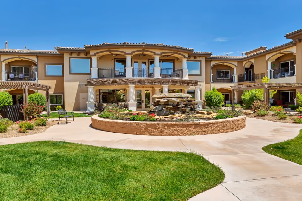 Two-story Mediterranean-style senior living building with a central courtyard fountain, walkways, balconies, and landscaping under a clear blue sky.