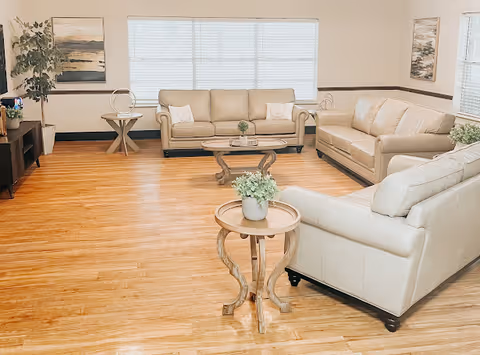 Bright communal living room featuring three beige sofas arranged around wooden coffee and side tables with potted plants and large windows.