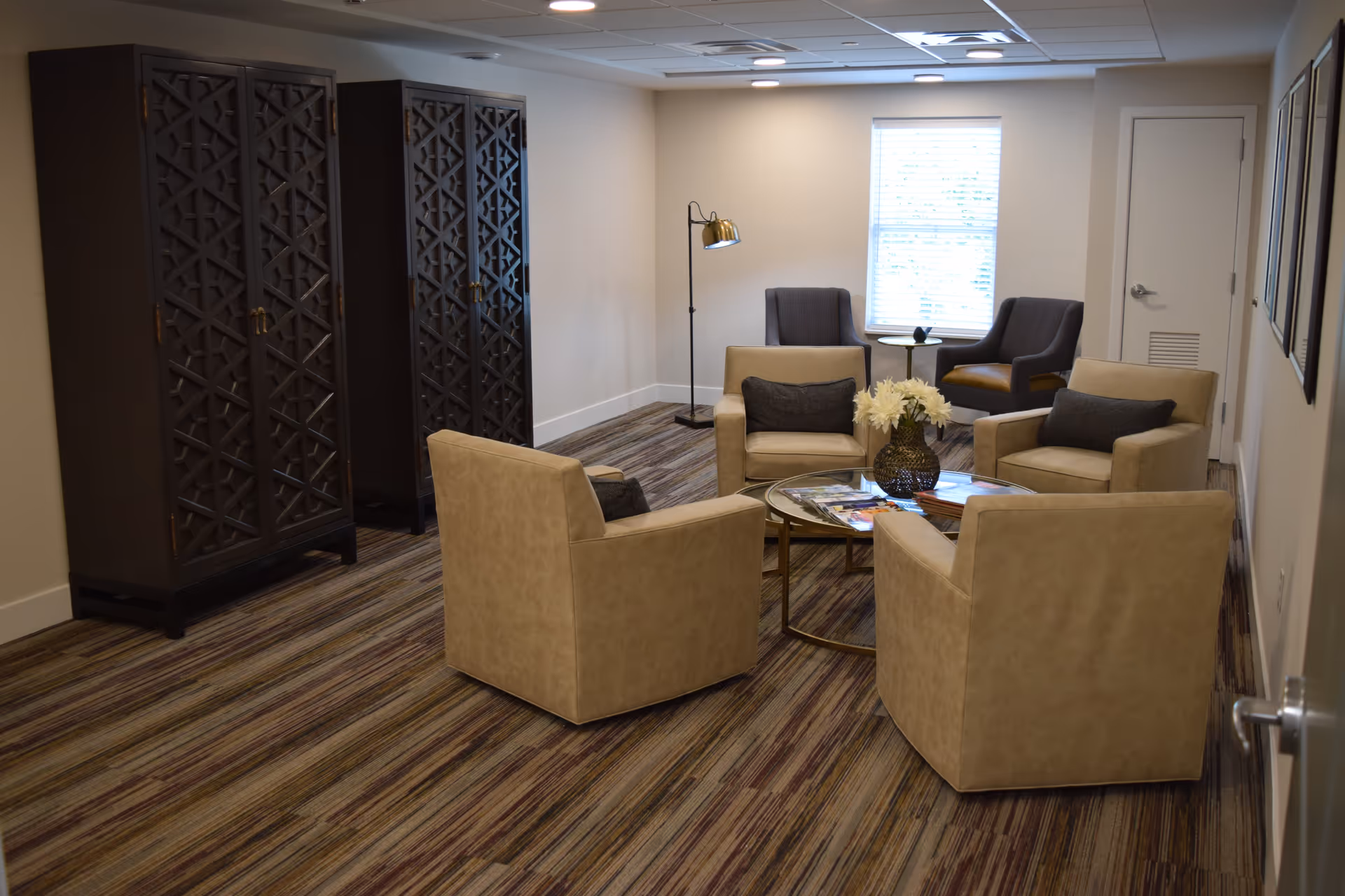 Small sitting room with four beige armchairs arranged around a glass coffee table, two additional chairs by a window, decorative cabinets along the left wall, and striped carpeting.