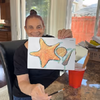An elderly person sitting at a table indoors, smiling and holding up a painting of three seashells including a starfish, a scallop shell, and a conch shell. There is a red cup and some art supplies on the table, with a window and a red fence visible in the background.