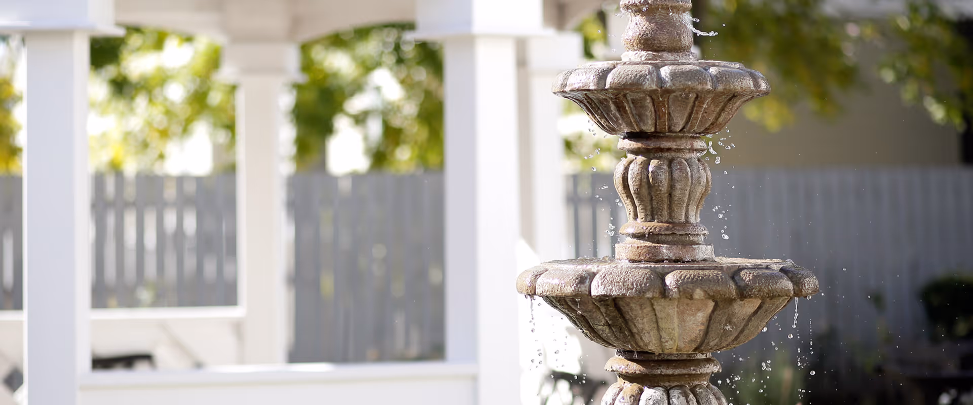 Stone tiered water fountain in a courtyard with pergola columns and a fence in the background.