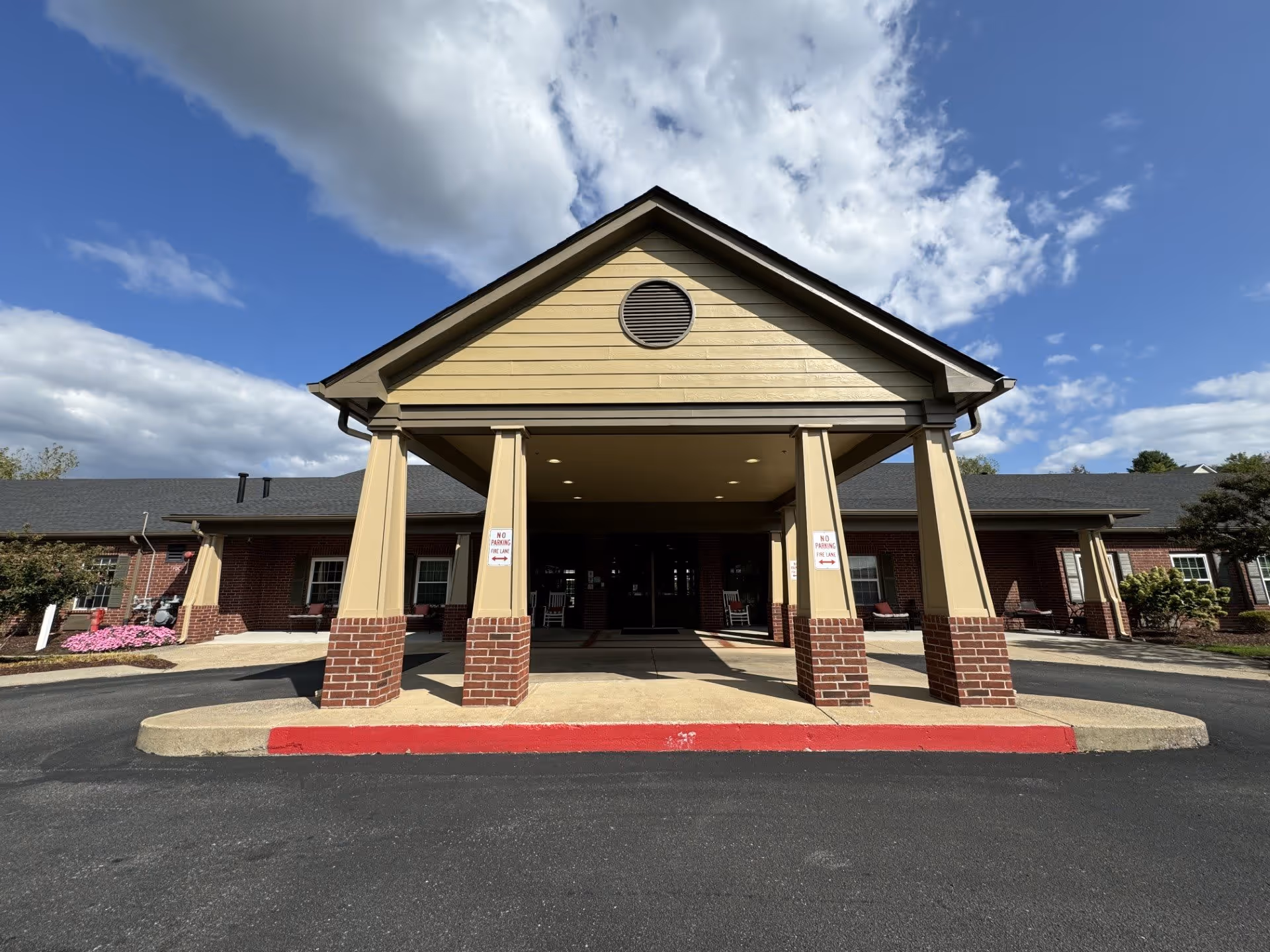 Front exterior view of a single-story building with a covered entrance supported by four columns. The building has a brick and beige siding facade, with a circular vent near the roof peak. There are signs on the columns indicating no parking in the fire lane. The sky is partly cloudy with blue patches visible.