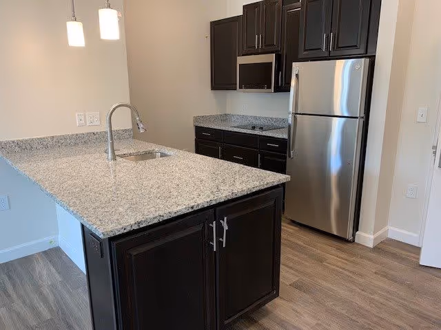 Modern kitchen with dark wood cabinets, a granite countertop island with a built-in sink and faucet, stainless steel refrigerator and microwave, and pendant lights hanging above the island. The floor is wood-style laminate.