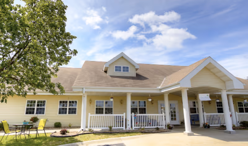 Exterior view of a single-story yellow senior living facility building with a covered porch supported by white columns, outdoor seating with a table and chairs on the left, and a large tree providing shade. The sky is blue with scattered clouds.