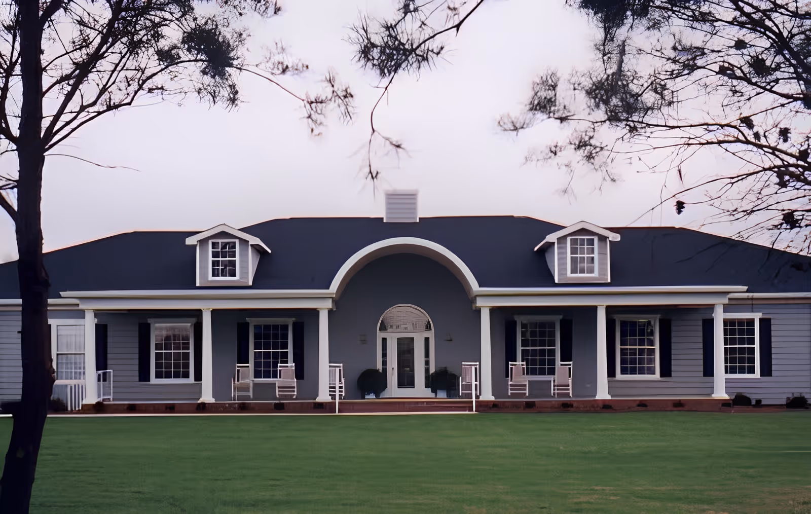 Front exterior view of a single-story senior living facility building with a dark roof, white columns, and multiple windows. There is a large arched entrance in the center with a glass door, and several rocking chairs are placed on the porch. Trees frame the image on the left and right sides, and a well-maintained green lawn is in the foreground.