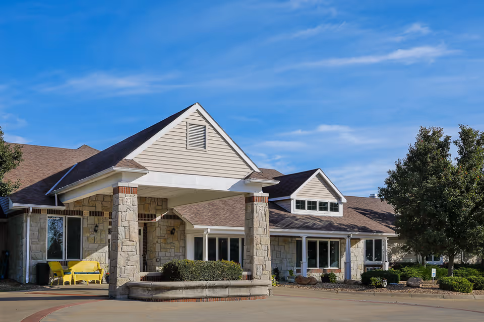 Stone-faced assisted living building front with a covered entrance, yellow seating, and landscaped shrubs under a blue sky.