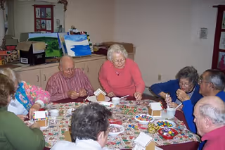 A group of elderly people sitting around a table engaged in decorating gingerbread houses with various colorful candies and icing in a cozy room.