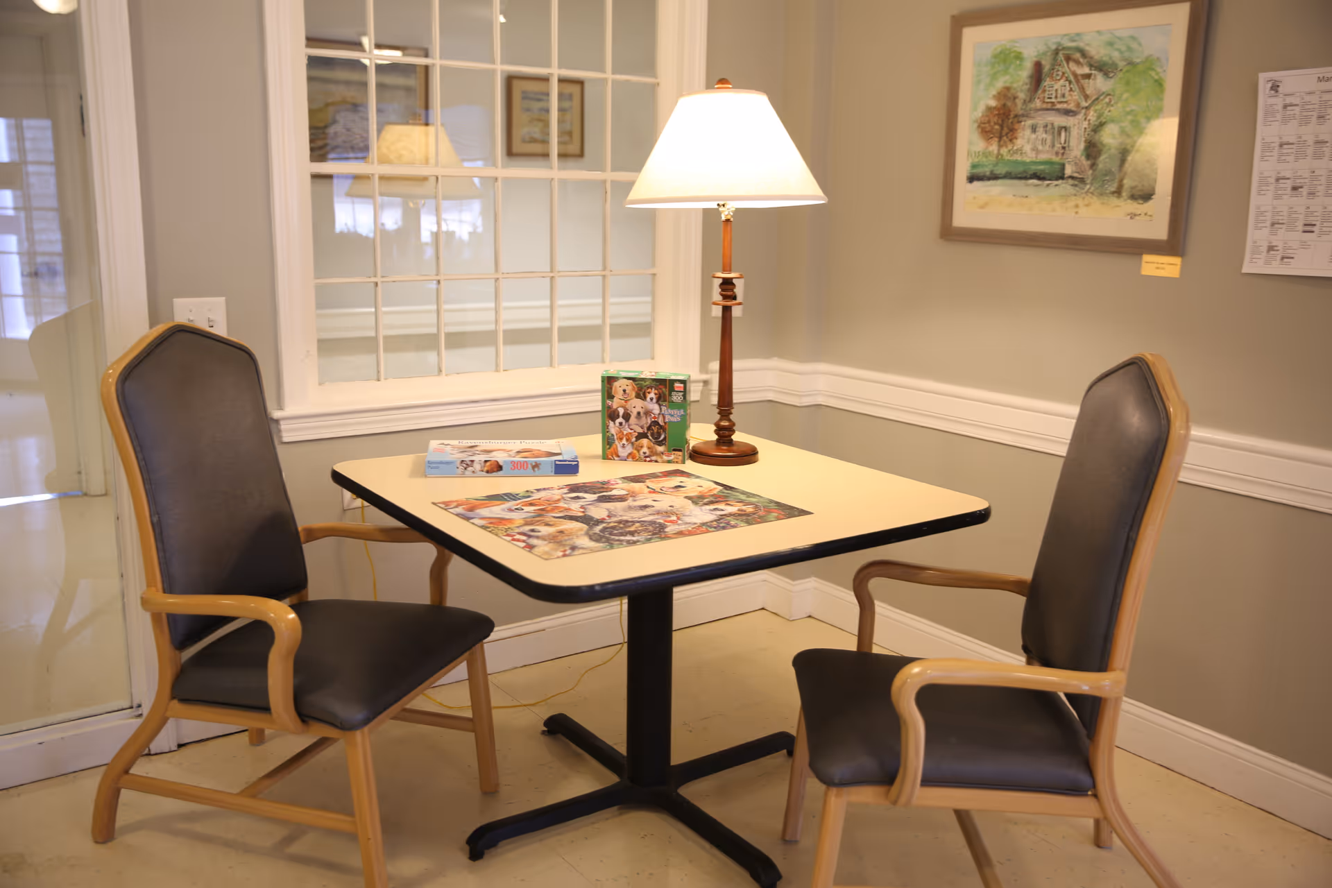 A small square table with a lamp and a partially completed dog-themed puzzle on it, flanked by two wooden chairs with black cushions. The room has beige walls with white trim, a framed painting, and a window with multiple panes behind the table.