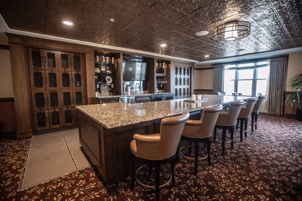 A spacious room with a large granite countertop island surrounded by six cushioned bar stools. The back wall features wooden cabinetry with glass shelves displaying glassware and decorative items, and a mounted flat-screen TV. The room has a patterned carpet, a decorative ceiling, and a large window with curtains letting in natural light.