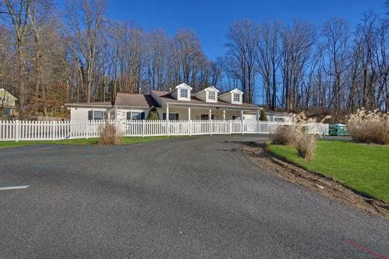 A single-story building with a white picket fence in front, surrounded by a paved driveway and green grass. Behind the building are leafless trees under a clear blue sky.