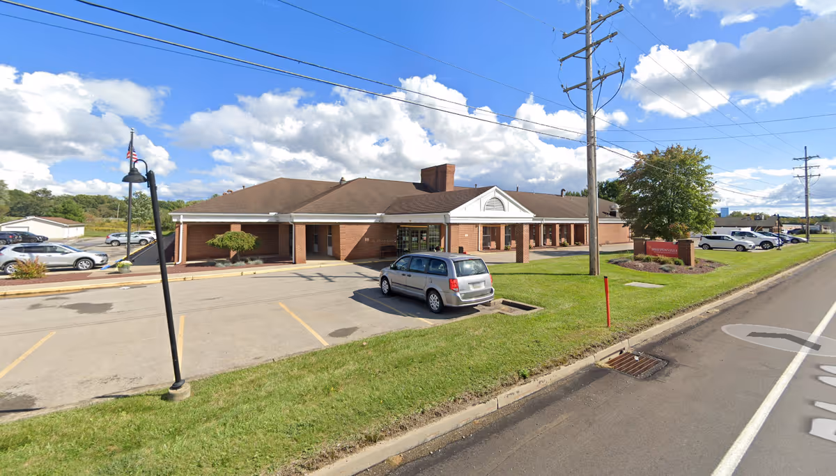Exterior view of Shippenville Nursing and Rehabilitation building with a parking lot in front, several parked cars, a grassy area, and a sign near the road. The sky is partly cloudy.