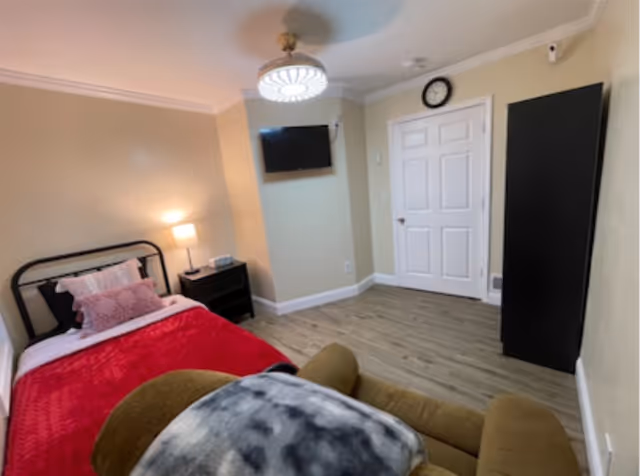 Small bedroom with a single bed, nightstand and lamp, wall-mounted TV, wardrobe, and recliner.