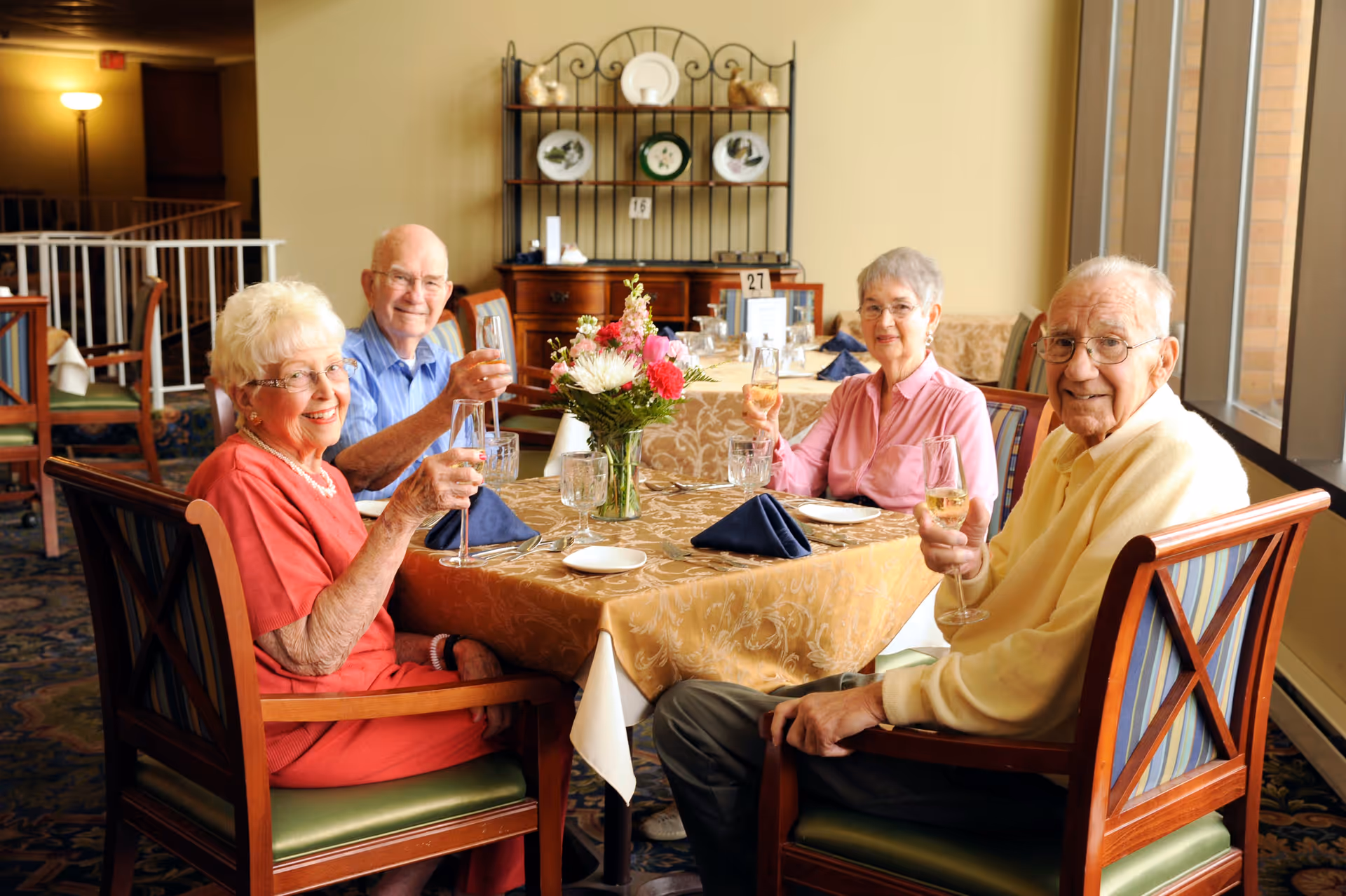 Four elderly people sitting around a dining table in a well-lit room, smiling and holding glasses of wine. The table is covered with a gold-patterned tablecloth, set with plates, glasses, and blue napkins. A vase with colorful flowers is in the center of the table. Behind them is a wooden cabinet with decorative plates and other items.