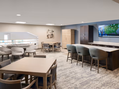 A spacious, well-lit common area with multiple seating options including a rectangular table with chairs, a round table with cushioned chairs, and a kitchen island with bar stools. A large flat-screen TV is mounted on the wall above a counter with cabinets. The room has a modern and comfortable design with carpeted floors and recessed lighting.