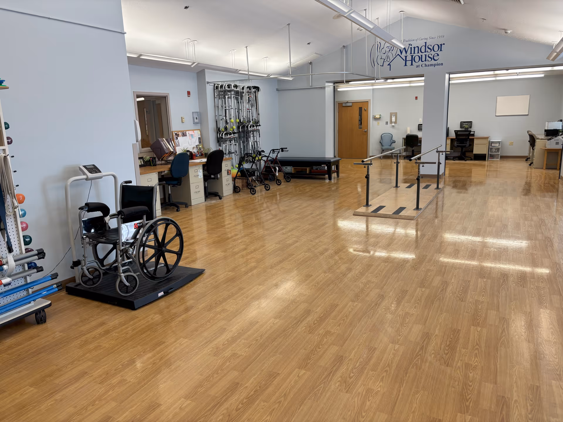 A spacious rehabilitation or physical therapy room with wooden flooring, parallel bars for walking exercises, a wheelchair on a platform, exercise equipment, walkers, and desks with chairs along the walls.